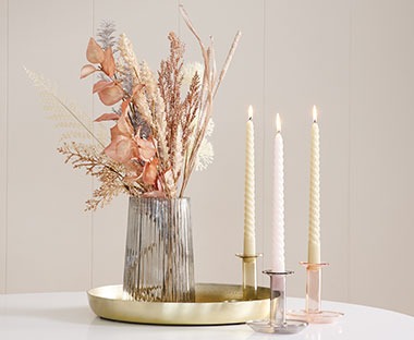Штучні рослини у вазі, яка стоїть на золотій таці, поруч три свічки у свічниках