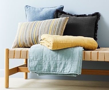 Два стьобані пледи синього та жовтого кольору, синя подушка і подушка кольору хакі, перед ними лежить подушка у різноколірну смужку