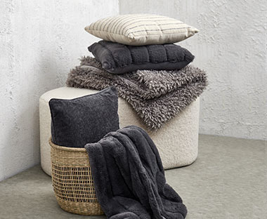 Декоративні подушки біло-сірих відтінків на пуфі та в плетеному кошику