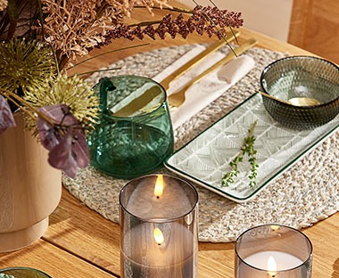 Обідній стіл сервірований свічками, скляний зеленим кухлем, мискою та тарілкою, які стоять на підставці з маісового волокна