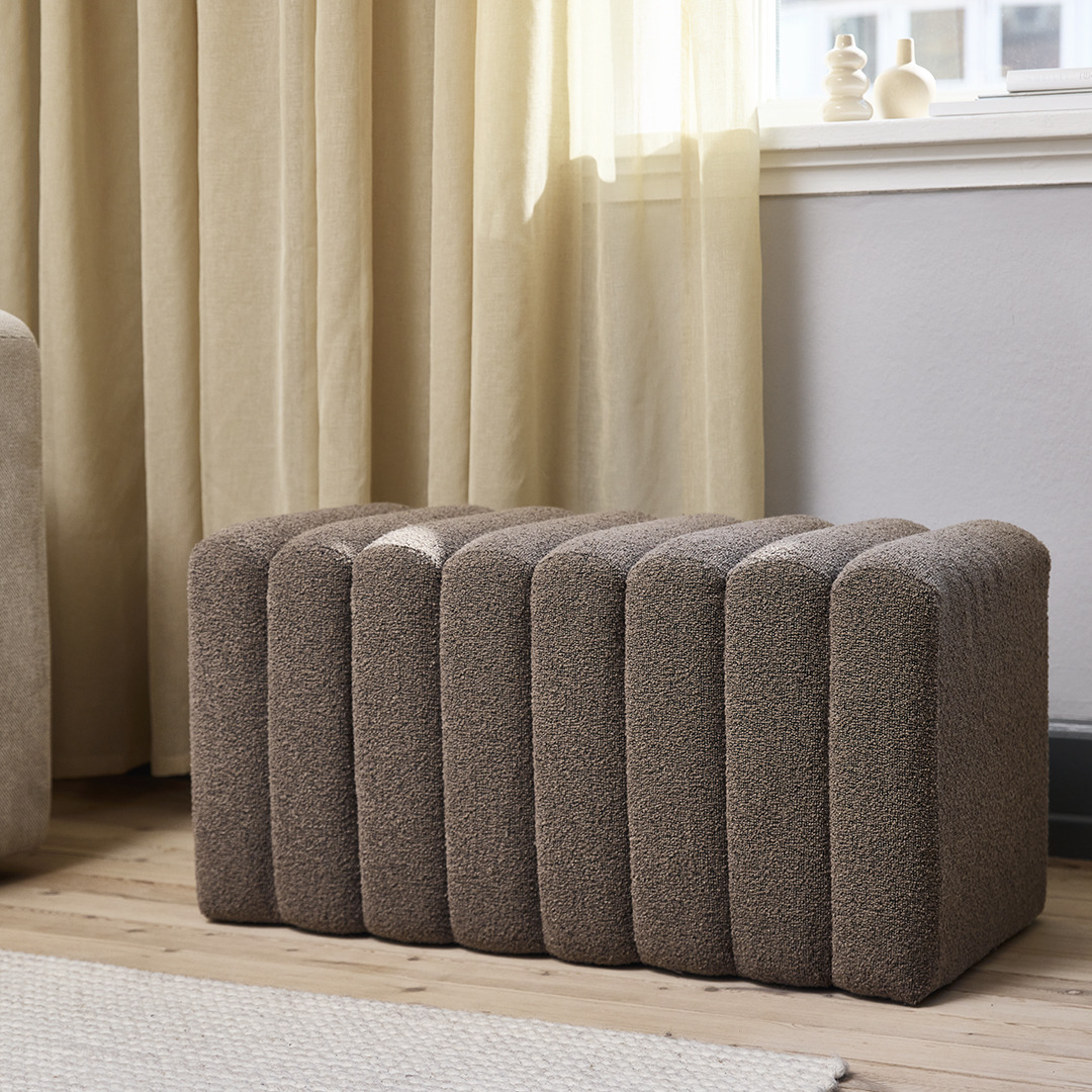 Банкетка оббита коричневою цупкою тканиною біля вікна зі шторами