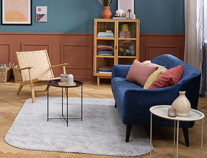 Синій диван з кольоровими подушками, плетене крісло та кавовий столик на сірому килимі у вітальні