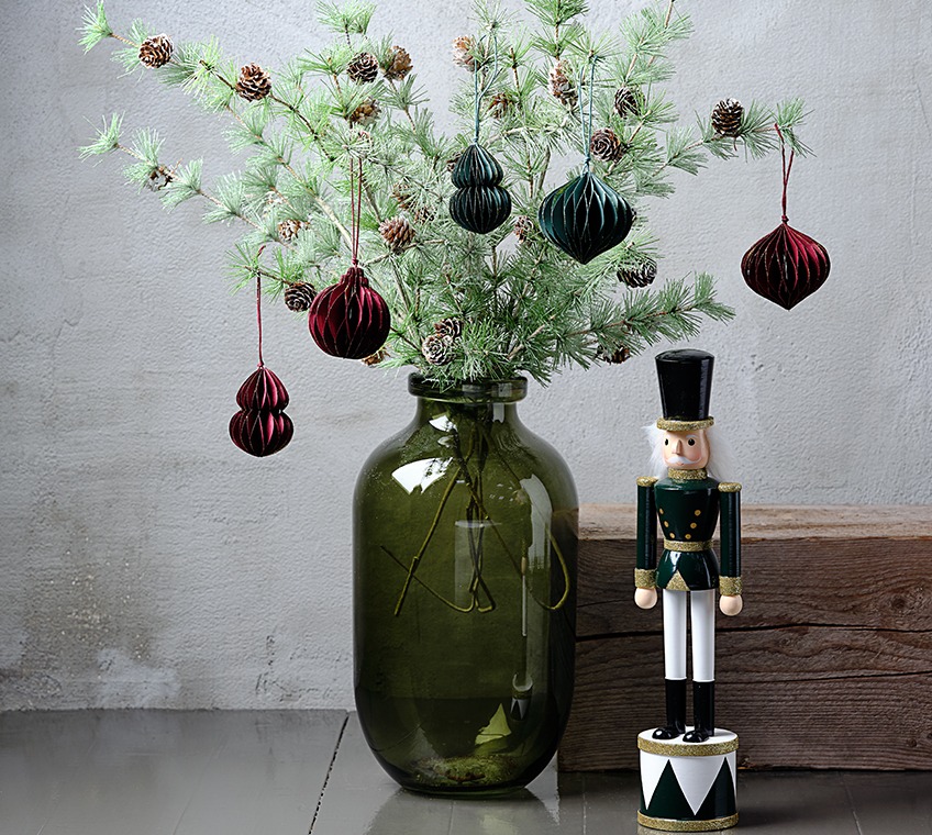 Green glass vase filled with artificial twigs with pinecones, decorated with Christmas ornaments in green and red. Beside it a Christmas figurine 