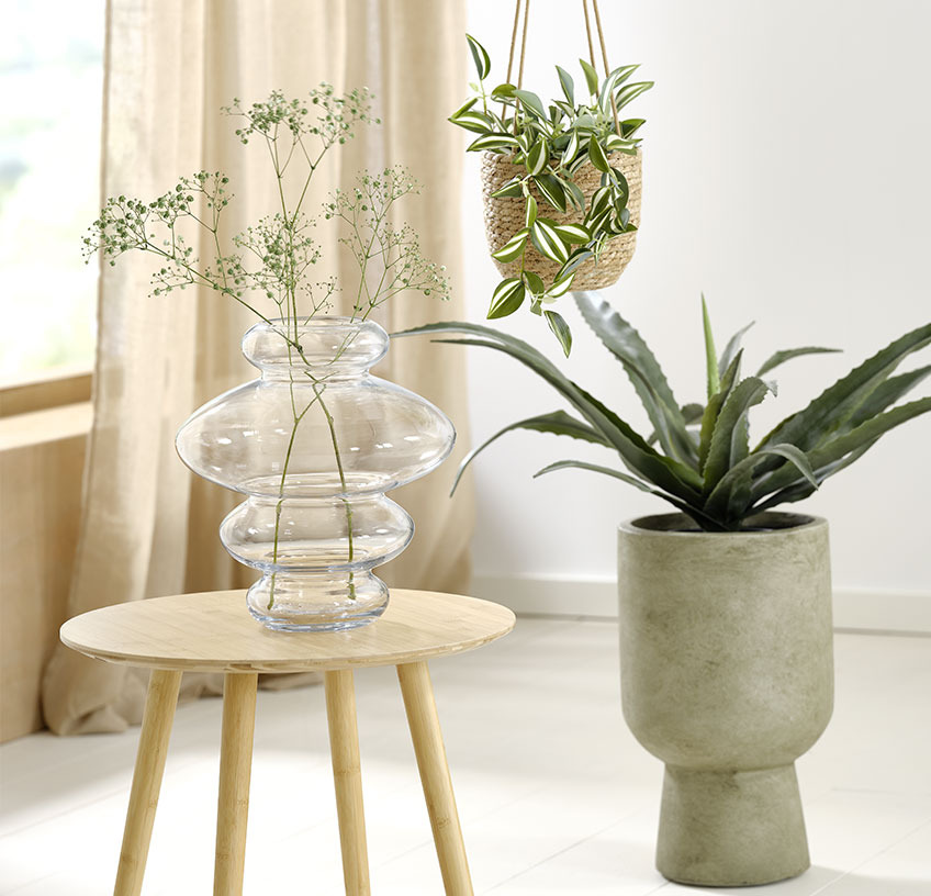 Скляна ваза на столику, підвісний горщик і зелений горщик зі штучними рослинами
