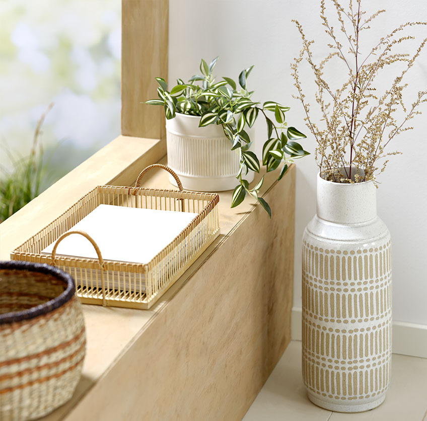 Висока ваза біля підвіконня з бамбуковою тацею і білим горщиком зі штучною рослиною